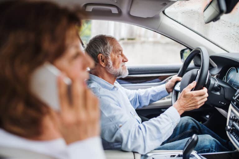 Et eldre par der mannen kjører bilen og damen snakker i telefonen.